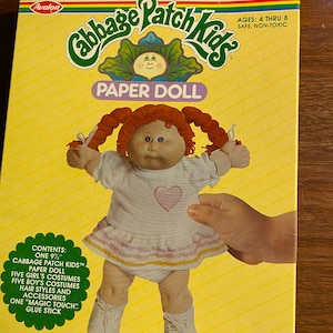 Op de afbeelding: Een Cabbage Patch Kids papierpoppenset met een 24 cm pop, vijf meisjesoutfits, vijf jongensoutfits, kapsels, accessoires en een lijmstok. De pop draagt een roze-wit gestreepte jurk met een roze hart op de voorkant.