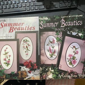Puede incluir: Dos libros de patrones de punto de cruz titulados "Summer Beauties" con colibríes y flores. El libro de la izquierda es el volumen uno y el libro de la derecha es el volumen dos.