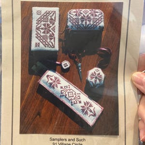 Puede incluir: Un surtido de artículos de punto de cruz, que incluyen un estuche para agujas, una caja pequeña y un colgante para tijeras, todos con un fondo blanco y un patrón geométrico burdeos. Los artículos están dispuestos sobre una superficie de madera.