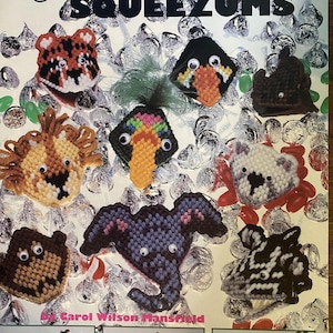 May include: A craft instruction booklet titled American School of Needlework plastic canvas SQUEEZUMS by Carol Wilson Mansfield. The cover features various animal-shaped plastic canvas projects with googly eyes, including a tiger, lion, and elephant.