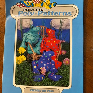 May include: A sewing pattern for a Freddie the Frog stuffed animal. The pattern is from Poly-Fil Poly-Patterns and is number D-210. The pattern shows three frogs, one sitting in a rocking chair, one sitting on the ground, and one standing. The frogs are all different colors and have polka dots.