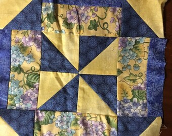 May include: A quilt block with a yellow and blue color scheme. The block is made up of nine squares, with a floral pattern in the center and a geometric pattern in the corners.