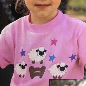 Op de afbeelding: Een roze T-shirt met een ontwerp van drie witte schapen, een bruin hek en blauwe en roze sterren. De schapen hebben zwarte ogen en neuzen.