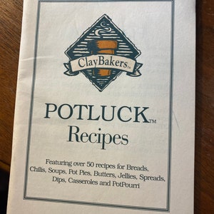 Puede incluir: Un libro de color beige con un borde negro y el título "POTLUCK Recipes" en texto negro. El libro presenta un logotipo con una taza de café marrón y el texto "Clay Bakers" en texto negro.