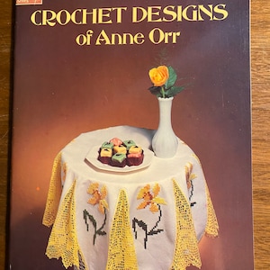May include: A book titled "Crochet Designs of Anne Orr" from the Dover Needlework Series. The cover features a white crocheted tablecloth with yellow daffodil embroidery, a vase with a yellow rose, and a plate of small, colorful candies.