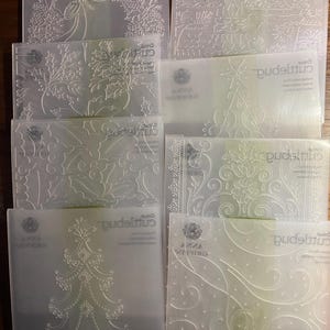 May include: A selection of clear plastic embossing folders featuring festive Christmas designs. The designs include a wreath, holly, a Christmas tree, and decorative patterns. These folders are for crafting and scrapbooking projects.