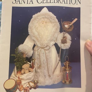 May include: A pattern titled "SANTA CELEBRATION" featuring a plush Santa Claus figure with a white coat, holding a bird on a branch. A sack of toys, a drum, and a lantern are also visible. The pattern is for a 21" St. Nicholas.