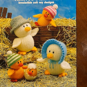 Può includere: Una copertina di libro che presenta una collezione di giocattoli di animali a maglia. La copertina mostra cinque animali a maglia: un pulcino, un'anatra, un pulcino in un uovo e due anatre. Il titolo del libro è "Jean Greenhowe's Knitted Animals" e il sottotitolo è "Irresistible soft toy designs". La copertina del libro include anche il testo "Double Knitting Yarn" e "USA: Knitting Worsted Weight".