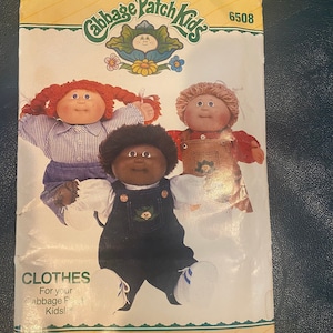 May include: A vintage sewing pattern for Cabbage Patch Kids clothes. The pattern is for making clothes for the dolls, and features three dolls on the front of the package. The pattern is from Butterick and has the number 6508 printed on it.