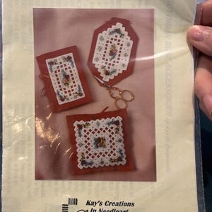 Op de afbeelding: Een pakket met de titel "Victorian Rose Accessories" met Hardanger en zijden lint. Het pakket toont drie rode stoffen stukken met witte kruissteekontwerpen en bloemaccenten. Een gouden schaar is ook zichtbaar.