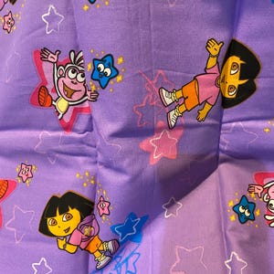 May include: Purple fabric featuring cartoon characters Dora the Explorer and Boots, along with blue, pink, and white stars. The fabric has a repeating pattern of the characters in various poses.