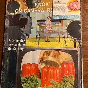 以下が含まれることがあります： 「KNOX ON-CAMERA RECIPES」というタイトルと「A completely new guide to Gel-Cookery」というサブタイトルが付いたヴィンテージの料理本の表紙。表紙には、キッチンにいる女性と、赤いゼラチンデザートが表示されたテレビ画面が描かれています。