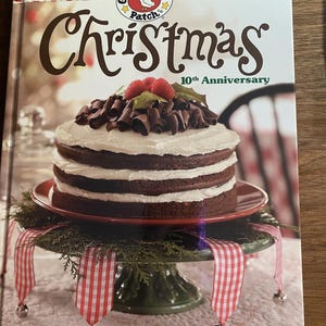 Puede incluir: Un libro de cocina de tapa dura titulado Gooseberry Patch Christmas 10th Anniversary que muestra un pastel de chocolate de tres capas con glaseado blanco, virutas de chocolate y frambuesas sobre un soporte verde con cintas rojas y blancas.