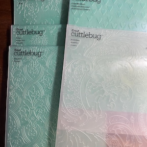Może przedstawiać: Kilka folderów do wytłaczania Cricut Cuttlebug. Foldery, w przezroczystych plastikowych opakowaniach, zawierają wzory takie jak serca i wzory kwiatowe. Widoczny jest tekst "Cricut cuttlebug". Foldery są w odcieniach zieleni i przezroczystości.