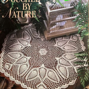 May include: A book cover with the title "Touched by Nature" and a large, white crocheted doily. The doily features a detailed, openwork design with pineapple motifs. A decorative birdcage and greenery are in the background. The book is about crocheted doilies.