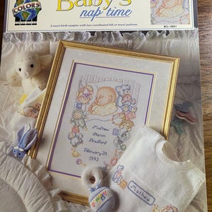 May include: A cross-stitch pattern titled "Baby's nap time" with a framed birth sampler, bib, and rattle. The sampler features a baby's name, birthdate, and illustrations of toys. The bib and rattle are white with embroidered details.