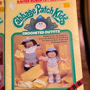 May include: A book cover with the title "Cabbage Patch Kids Crocheted Outfits". The cover features two Cabbage Patch Kids dolls, one wearing a blue and white striped outfit and the other wearing a pink and white outfit. The book is for crocheting clothing for Cabbage Patch Kids dolls.
