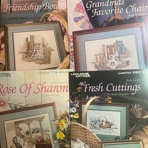 May include: A collection of framed cross-stitch patterns, including "Friendship Bouquet," "Grandma's Favorite Chair," "Rose of Sharon," and "Fresh Cuttings." Each features a detailed, colorful design with a wooden frame. The leaflets are by Paula Vaughan.