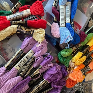 May include: A collection of colorful embroidery floss in various shades, including red, purple, blue, green, and yellow. The spools are neatly arranged, with labels visible, suggesting a craft or sewing project. The image showcases a variety of colors and textures.