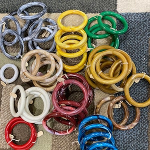 May include: A collection of colorful bangles in various shades, including blue, red, green, yellow, and brown. Some bangles have a marbled effect. The bangles are made of a glossy material and are arranged on a patterned surface.