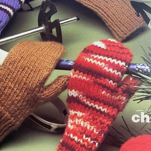 May include: A close-up of a pair of red and white striped knitted mittens. The mittens are made with a chunky yarn and have a textured knit. The mittens are lying on a green surface with a brown knitted mitten and a ski pole in the background.