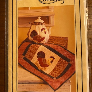May include: A package for a tea cosy and place mats set. The set features a rooster design. The place mats measure 33 cm by 51 cm. The package is labelled "Pearl Louise Designs" and "#374 Rise & Shine".