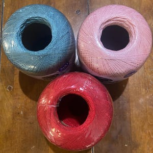 May include: Three cylindrical balls of crochet thread arranged in a triangle formation. The top left ball is teal, the top right ball is light pink, and the bottom ball is red. Each ball features a central hollow core and a partial paper label.