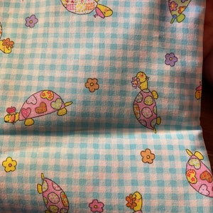 Puede incluir: Tela de vichy azul y blanca con un patrón repetido de tortugas y flores de colores. Las tortugas tienen caparazones rosas, amarillos y verdes con flores en la cabeza.
