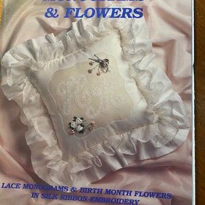 Könnte beinhalten: Ein Buch mit dem Titel "Monogramme & Blumen" mit einem weißen Kissen, das mit dem Wort "Home" in einer Schreibschrift bestickt ist. Das Kissen ist mit Seidenbandblumen und Spitzenbesatz verziert. Der Buchumschlag hat einen rosa Hintergrund und den Text "Spitzenmonogramme & Geburtsmonatsblumen in Seidenband-Stickerei von Judith Marquis".