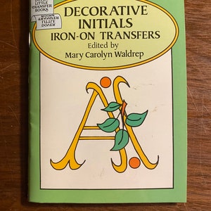 Può includere: Un libro intitolato "Iniziali decorative Trasferibili a caldo" a cura di Mary Carolyn Waldrep. La copertina presenta uno sfondo verde con una lettera A gialla decorata con foglie verdi e cerchi arancioni.