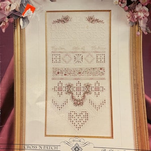 May include: A framed cross-stitch sampler with a gold-colored frame. The sampler features various stitched patterns, including floral designs, geometric shapes, and a heart. The text "Handen Malo of Hindn000" is visible. The frame is decorated with pink and white flowers.