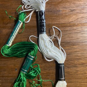 Könnte beinhalten: Drei Spulen Stickgarn in Weiß und Grün. Eine weiße Spule hat ein schwarz-silbernes Etikett mit dem Text "INGER MAR". Zwei grüne Spulen haben Etiketten mit Text wie "COULEURS SOLIDES COLORFAST" und "8m-8.7y".