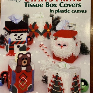 以下が含まれることがあります： クリスマスのテーマで飾られたティッシュボックスカバー4つが載っているクラフト本の表紙。カバーはプラスチックキャンバスで作られており、雪だるま、ポインセチア、サンタクロース、クリスマスツリーが描かれています。本のタイトルは「Christmas Tissue Box Covers in plastic canvas」で、著者はSue Penrodです。