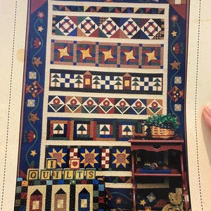 Puede incluir: Un patrón de edredón colorido con varias formas geométricas y diseños de estrellas. El edredón presenta una mezcla de bloques azules, rojos, amarillos y verdes. El texto "I QUILTS" se muestra en bloques de letras de madera. Se ve una pequeña estantería de madera con un oso de peluche.