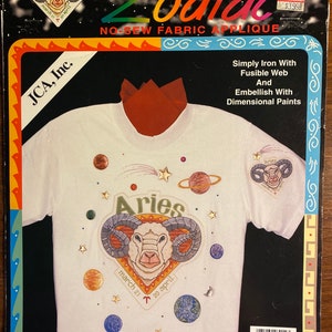 May include: A white t-shirt with a colorful iron-on applique of the zodiac sign Aries. The applique features a ram's head with a white face and gray horns. The background of the applique is a red circle with the words "Aries", "March 21", and "20 April" written in black. The t-shirt is surrounded by a blue and white border.
