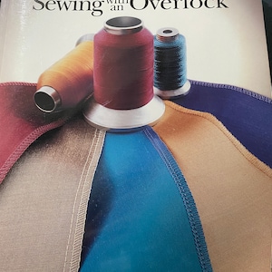 Pode incluir: Um livro intitulado "Costurar com uma Overlock" da Biblioteca de Referência de Costura Singer. A capa apresenta três carretéis de linha em vermelho, laranja e azul, e um close-up de costuras de tecido.