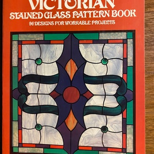 May include: A softcover book titled ED SIBBETT, JR. VICTORIAN STAINED GLASS PATTERN BOOK 96 DESIGNS FOR WORKABLE PROJECTS. The cover features a red background and a central illustration of a geometric stained glass window with blue, purple, and green glass panels.