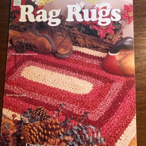 May include: A book cover with the title "Quick & Easy Rag Rugs" in large, bold letters. The cover features a crocheted rug in shades of red and white, with a pair of brown leather boots and a wooden duck figurine. The text "Designs by Nazanin Fard" and "Rustic Log Cabin" are also visible.