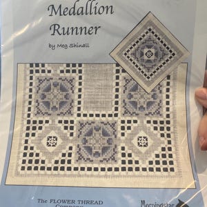 Op de afbeelding: Een pakket met een kruissteekpatroon voor een "Medallion Runner" van Meg Shinall. Het ontwerp bevat geometrische vormen in blauw- en grijstinten. Het pakket toont ook de logo's van de Flower Thread Company en de Morningstar Embroidery Group.