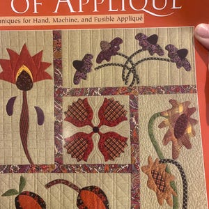 Peut inclure: Couverture de livre intitulée "The Easy Art of Appliqué" présentant un motif matelassé. Les blocs de courtepointe présentent des motifs floraux et abstraits dans des tons de rouge, marron et vert sur fond beige. Le design comprend diverses techniques d'appliqué.