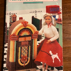 Puede incluir: Un folleto de patrones de manualidades para una tienda de soda de muñecas de moda. La portada presenta una muñeca con una falda roja con un diseño de caniche blanco, de pie junto a una máquina de discos colorida. El título del folleto es "Fashion Doll Soda Shop" y el número 87D25 está impreso en la esquina superior derecha.