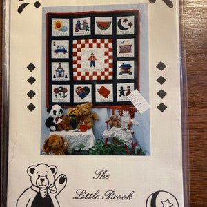 May include: A white background with a black and white illustration of a teddy bear wearing a bow tie. The text "The Little Brook Collection" is below the bear. The image also includes a quilt pattern with a variety of colourful squares and a black and white checkered square in the centre. The quilt pattern is surrounded by a white border.