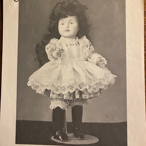 Puede incluir: Fotografía en blanco y negro de una muñeca de reproducción llamada "Gretchen I" (KR 114 POUTY) que lleva un vestido blanco con ribete de encaje y un delantal blanco. La muñeca mide 30,5 cm de alto y está de pie sobre un soporte. El patrón de la muñeca es de Shirley Augustine. SA-6 Reproduction Doll "Gretchen I" (KR 114 POUTY) (12") Doll, Dress and Pattern by: Shirley Augustine 261 West Commercial Street, Pomona, CA 91768 Pattern Copyright 1974 (Revised 1977) Shirley Augustine Pattern $3.00