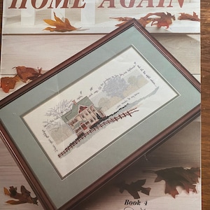 May include: A framed cross-stitch of a house with a red roof and a fence, with the words "Home Again" at the top. The frame is brown, and the matting is a light green. The book cover has autumn leaves.
