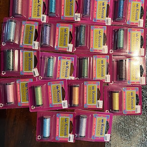 May include: A collection of 18 spools of Kreinik metallic embroidery floss in various colors, including gold, silver, pink, blue, green, and brown. Each spool is labeled with the brand name "Kreinik" and the word "Metallic".