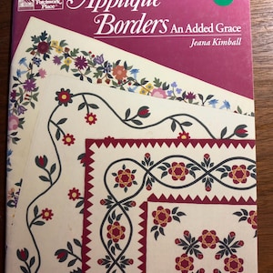 Può includere: Un libro con copertina flessibile intitolato Appliqué Borders An Added Grace di Jeana Kimball. La copertina presenta uno sfondo magenta intenso con un logo bianco e tre illustrazioni sovrapposte di tessuto color crema con disegni floreali per bordi di trapunte.