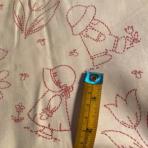 May include: White fabric with red embroidered designs of a girl in a hat, flowers, and tulips. A measuring tape is placed on the fabric to show the size.