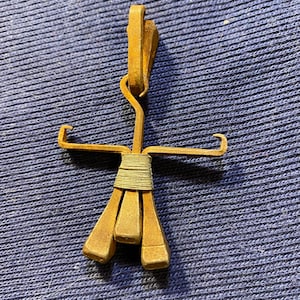May include: A gold-colored metal pendant shaped like a stick figure with a brown leather strap around the waist. The pendant is made from repurposed horseshoes.