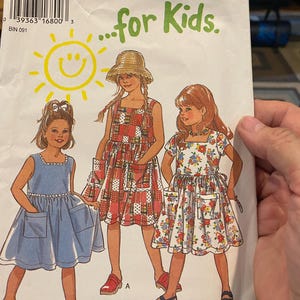 May include: A sewing pattern titled "NEW LOOK ...for Kids" featuring illustrations of three young girls in dresses. The dresses are in blue, red, and floral patterns. The pattern size is 3-8.