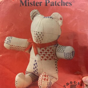 Puede incluir: Un oso de peluche llamado "Mister Patches" hecho de tela beige con retazos rojos, blancos y azules. El oso tiene un lazo rojo y está sobre un fondo rojo. El texto "Mister Patches" está en la parte superior.